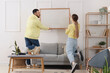 © Pixel-Shot - Young couple hanging blank frame on light wall at home, back view