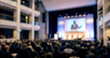 © AJI EGY TAMA - Fully blurred global economic forum hall with executive speaker and audience silhouettes.