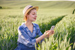 © sofiko14 - A woman in a straw hat and overalls uses her mobile phone while standing in a lush green wheat field during golden hour