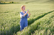 © sofiko14 - A woman in overalls and a straw hat stands in a lush green wheat field, examining a stalk while holding her smartphone to take a picture or record data