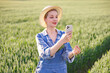 © sofiko14 - A woman in overalls and a straw hat examines a wheat stalk while holding a smartphone in a lush green field