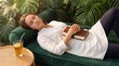 © sabda - Peaceful woman sleeping on a green velvet sofa with a book surrounded by lush plants.