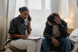 © Seventyfour - Black female young adult social worker sitting on bed, talking on phone and writing notes while Black male teenager client sitting nearby, holding head during counseling session