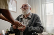 © Seventyfour - Senior Caucasian man with beard sitting in wheelchair receiving cup from Black woman social worker, looking up with attentive expression in indoor setting