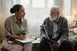 © Seventyfour - Young adult Black woman social worker holding clipboard talking with senior Caucasian man with beard sitting on bed, holding walking cane and tablet during home visit