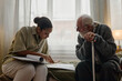 © Seventyfour - Young adult Black woman social worker assisting senior Caucasian man with cane by reviewing documents together on bed, both focused on paperwork in residential setting