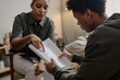 © Seventyfour - Young adult Black woman explaining documents to young zoomer during social work consultation, both seated indoors, woman gesturing toward paperwork, man attentively reading
