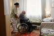 © Seventyfour - Black woman assisting senior Caucasian man with disability in wheelchair moving near bed in residential room, social worker supporting elderly client during daily routine