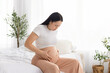 © Anastasiya - Pregnant woman sitting on bed holding belly and feeling contractions discomfort. Pregnancy health issue concept including abdominal pain, prenatal care and maternal wellbeing