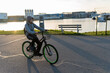 © CatalnIlie - Young man wearing a hoodie cycling an e-bike on an asphalt path next to a river with industrial buildings in background