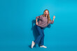 © deagreez - Young woman dancing in a cozy sweater and jeans against blue background smiling and joyful