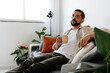 © kleberpicui - Man sitting on a sofa holding a cup of coffee and looking thoughtful in a bright living room, representing calm moments, relaxation and everyday home routine.