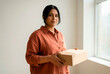 © designX - South Asian woman holding a cardboard box in a bright minimalist room while looking at the camera during a house relocation