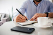 © kleberpicui - Close-up of a man writing notes in a notebook while working at a desk with laptop, coffee and smartphone, representing planning, productivity and remote work.