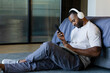 © Wavebreak Media - African American man sitting on rug by low bed wearing headphones, checking phone, writing notebook