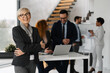 © Novak - Successful blonde woman professional standing in office with coworkers behind her.