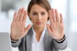 © whitestorm - A businesswoman showing stop gesture with her hands, preventing a further action, expressing negation and warning, wearing a gray jacket and sitting behind a laptop at the office.