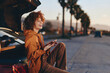 © SHOTPRIME STUDIO - Young woman with curly hair wearing rainbow sweater sits on car trunk smiling and holding smartphone outdoors on sunny day with palm trees and road in background.