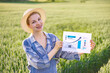 © sofiko14 - A smiling woman in a straw hat and overalls stands in a green field, holding a paper with a rising graph and statistics