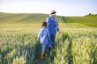 © sofiko14 - A farmer wearing overalls and a hat walks with a young child through a lush green wheat field under a clear sky
