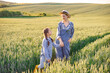 © sofiko14 - A woman and a young girl in denim overalls stand holding hands in a vast wheat field during golden hour, sharing a tender moment