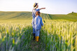 © sofiko14 - A woman in a straw hat and overalls points towards the horizon while walking with a young girl through a lush green wheat field at sunset
