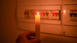 © Nanci - Hand holding a lit candle, providing light in front of an electrical panel with circuit breakers, illustrating a power cut and the absence of electricity in a home