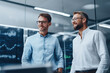 © sam richter - Two smiling businessmen stand together in modern office with financial screens.