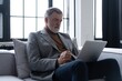 © opolja - Happy smiling mature man wearing suit looking at laptop using computer watching webinar sitting at sofa.
