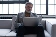 © opolja - Happy smiling mature man wearing suit looking at laptop using computer watching webinar sitting at sofa.