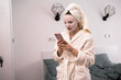 © Andrii Lysenko - young woman in cosmetic sheet mask on face with towel on head using mobile phone at home. beauty, skincare and cosmetic product concept