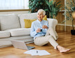 © Lumos sp - Portrait of a senior woman working on laptop drinking coffee dring and  sitting on the floor at home