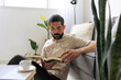© kleberpicui - Adult man relaxing on sofa reading book at home with coffee nearby. Calm lifestyle moment representing mindfulness, rest and slow living in modern bright interior.