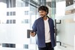 © Tetiana - An Indian smiling man holds a phone in his hands, opens the door and enters the office premises