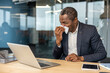 © Liubomir - Mature businessman experiencing seasonal allergies or cold symptoms, sneezing into a tissue while working at a laptop in a contemporary office environment, prioritizing health while at work