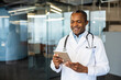© Liubomir - African american male doctor in a white lab coat with stethoscope smiling while using a digital tablet modern telemedicine care, professional healthcare consultation in clinic