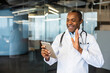 © Liubomir - Male doctor wearing a lab coat and stethoscope, smiling and waving while holding a tablet computer, providing online medical consultation in an office environment