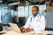 © Liubomir - Experienced physician wearing a white lab coat and stethoscope, sitting at a desk and typing on a laptop, providing telemedicine services from a contemporary healthcare clinic