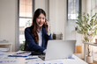 © David - Happy Asian businesswoman talking on smartphone while looking at laptop screen in modern office