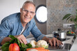 © luciano - Smiling mature man searching for new recipes on his laptop while cooking healthy food at home in a modern kitchen, preparing vegetables. Concept of nutrition, active lifestyle, and healthy eating