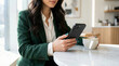 © illustratica - Woman in green blazer using smartphone while holding a cup of coffee at a cafe