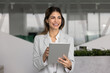 © fizkes - Confident smiling Hispanic female professional project manager executive in formal suit distracted from digital tablet screen looking forward planning optimistic business strategy and goals in office