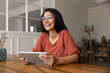 © fizkes - Joyful Latina woman holding tablet seated at table indoors, looking away with smile, enjoy digital communication with family, reading pleasant news, chatting with friend through modern application