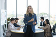 © eakgrungenerd - Confident businesswomen holding clipboard standing in office during team meeting, professional leader reviewing documents while coworkers, concept of leadership, management and teamwork.