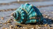 © blu - Close-up of a colorful blue seashell on the beach with small pebbles and the ocean in the background under bright sunlight, perfect for summer vacation themes.