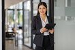 © David - A poised businesswoman, elegantly dressed, stands near an office entrance with a tablet, projecting confidence and readiness to engage with clients and colleagues alike.