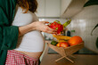 © wifesun - Pregnant woman standing in her kitchen, selecting a dragon fruit from a wooden bowl filled with various fresh fruits, emphasizing healthy eating habits and prenatal nutrition