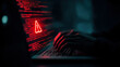 © Alina Tymofieieva - Cybersecurity warning shows hands typing on a keyboard in a dark room with red warning sign on screen indicating a potential threat