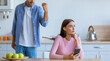 © Prostock-studio - A man is showing frustration in a kitchen while a woman sits at a table with her phone. Green apples are on the table. The atmosphere shows tension between the couple.