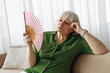 © Anastasiya - Senior woman cooling herself with handheld fan at home living room. Mature caucasian female feeling hot and uncomfortable, illustrating summer heat or menopause symptoms in retirement lifestyle